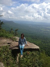 Portrait of smiling mid adult woman sitting cliff against cloudy sky