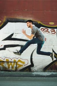 Side view of young man jumping against graffiti wall