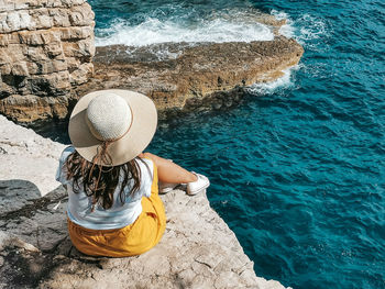 Young woman in yellow skirt sitting on edge of cliff above sea.