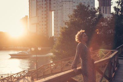 Woman leaning against railing while standing in city during sunset