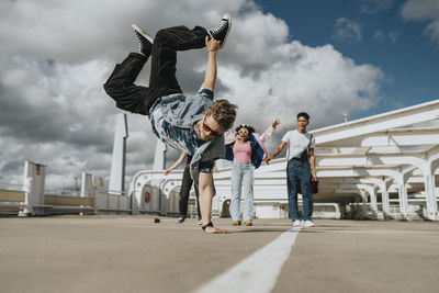 Athletic young man doing handstand while male and female friends cheering in parking lot under cloudy sky