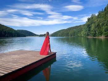 Woman wearing long red dress standing on a wooden pontoon near a lake surrounded by forest