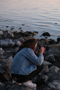 Rear view of girl taking photo sitting on rock at sea shore