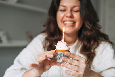Adult woman plus size body positive in white cozy knitted sweater with cupcake with candle in hands