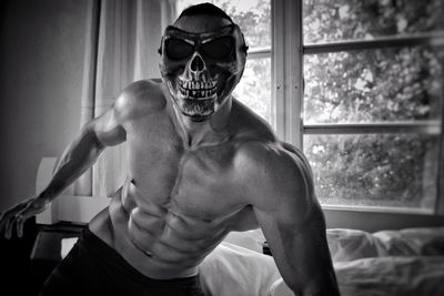 Muscular man wearing skull mask leaning against window at home