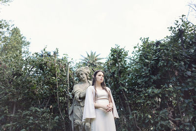 Thoughtful fairy elf young woman standing by sculpture and trees