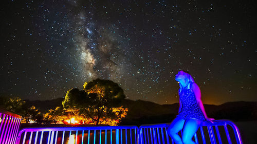 Low angle view of woman sitting on railing against milky way at night