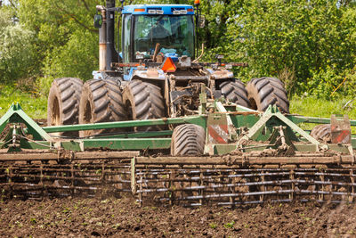 Blue tractor with double wheels pulling disc harrow with roller basket at hot sunny day