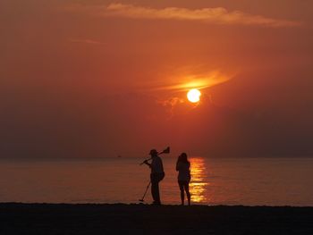 Silhouette people standing at beach against orange sky