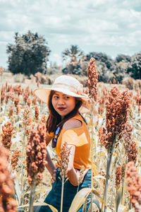 Side view of smiling young woman wearing hat while posing amidst plants against sky during sunny day