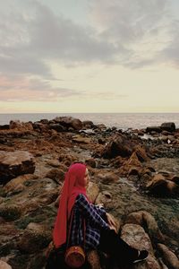 Woman sitting on rock at beach against sky during sunset