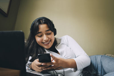 Smiling teenage girl using mobile phone and laptop while lying on bed at home