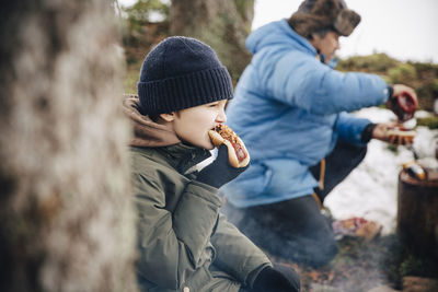 Boy in warm clothing eating hot dog while father preparing food