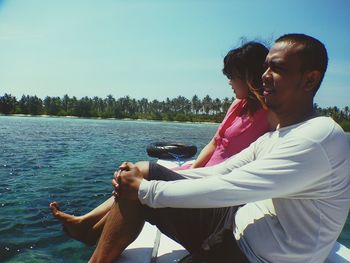 Side view of couple sitting on shore against clear sky