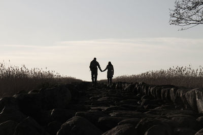 Rear view of silhouette couple holding hands while walking on rocks against sky during sunset