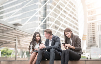 Business colleagues using smart phone while sitting against buildings in city