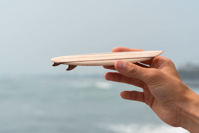 Cropped hand of person holding small surfboard against sky