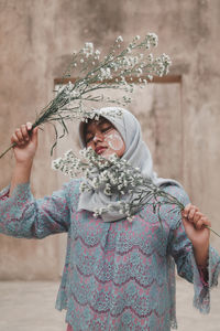 Woman wearing hijab holding flowers while standing against wall