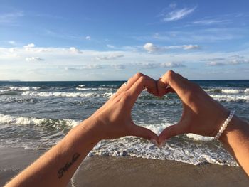 Cropped hand of woman making heart shape at beach against sky