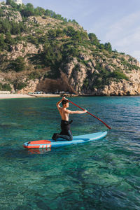 Back view of full body male surfer knelling on paddleboard with oar in hands while practicing sup surfing on turquoise seawater in sunny day