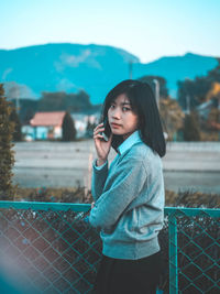 Portrait of young woman talking over mobile phone while standing by fence