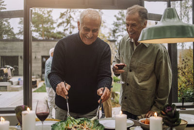 Happy mature man tossing salad while standing with male senior friend at dinner party