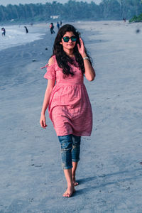 Full length portrait of young woman wearing sunglasses while walking on shore at beach