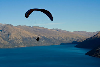 Person paragliding over lake and mountains against clear blue sky