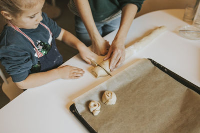 Mother holding cinnamon roll dough while daughter cutting with knife at table