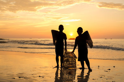 Silhouette boys holding birdcage while standing at beach against sky during sunset