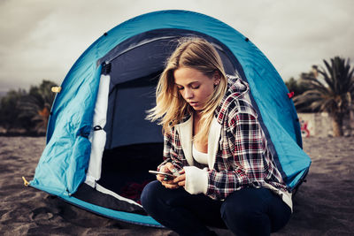 Young woman using mobile phone while crouching by tent
