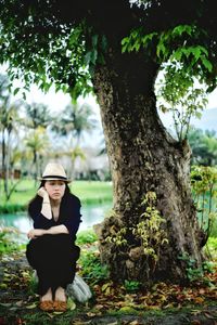 Full length of sad woman sitting by tree trunk