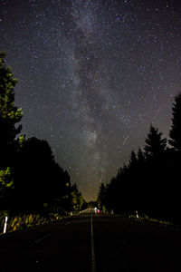 Empty road amidst trees against star field at night