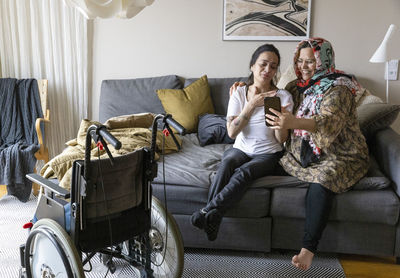 Woman with paraplegia doing video call through smart phone while sitting with mother on sofa at home