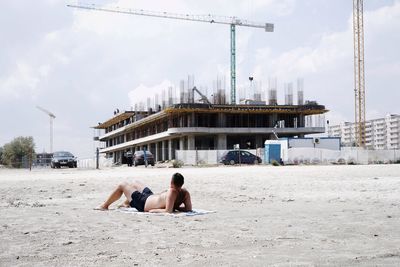 Shirtless man relaxing at beach against incomplete building in city