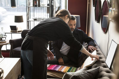 Rear view of young female owner showing fabric swatch to male customer sitting on sofa at shop