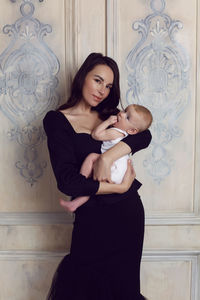 Mother in black dress holds a newborn boy in her arms at the window