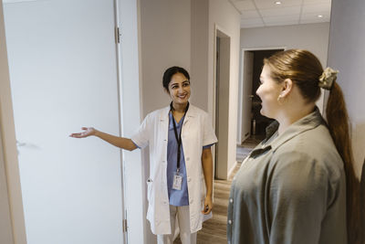 Smiling doctor assisting female patient towards medical examination room at clinic