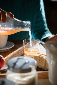 Unrecognizable female sitting at table in kitchen and pouring fresh juice from bottle in glass while having delicious domestic breakfast