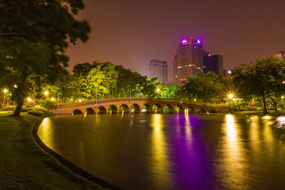 Illuminated modern buildings by river against sky at night