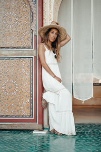 Portrait of young woman wearing hat standing by wall