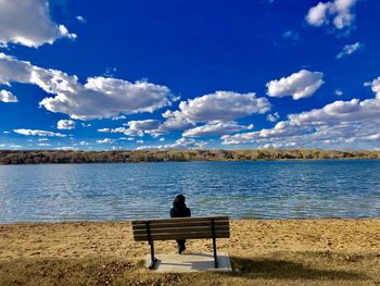 Rear view of man sitting on bench by lake against sky