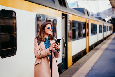 Woman using mobile phone while standing against train at platform