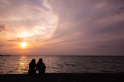 Silhouette couple sitting by sea against sky during sunset