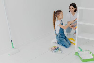 Full length of mother and daughter holding paint roller sitting at home