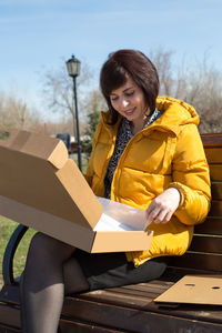 A happy brunette woman in a bright yellow jacket bench with a smile opens a box with a new laptop.