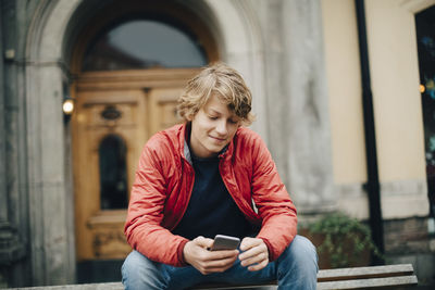 Smiling teenage boy using mobile phone while sitting on bench in city