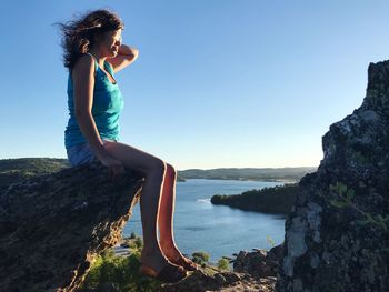 Side view of young woman on cliff at beach against clear blue sky