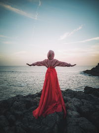 Rear view of girl with arms outstretched standing on rock at beach during sunset