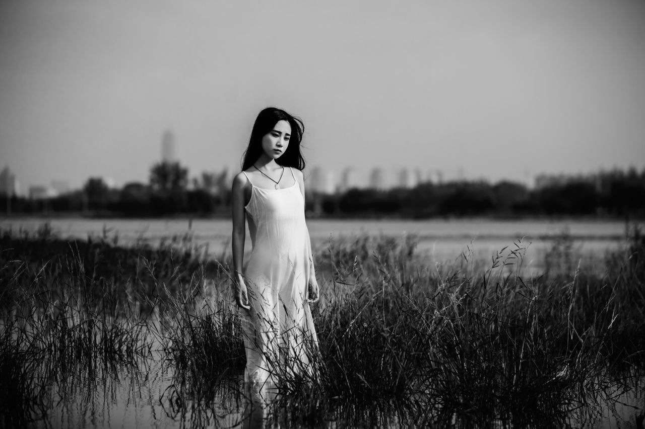 one person, field, real people, three quarter length, lifestyles, young adult, leisure activity, nature, young women, standing, grass, water, outdoors, lake, front view, beautiful woman, sky, plant, growth, beauty in nature, portrait, clear sky, scenics, looking at camera, full length, women, day